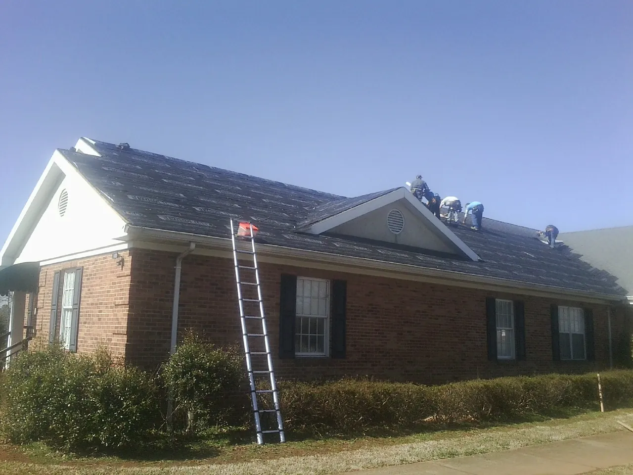 Crew tearing shingles off a brick ranch roof with a ladder set — Emergency Roof Repair work by our crew in Crookston
