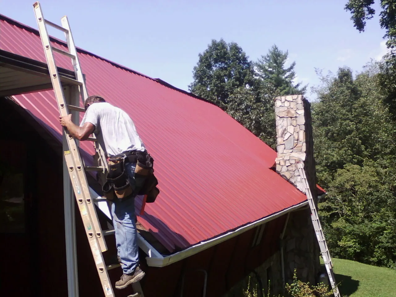 Roofer on a ladder beside a red metal roof and stone chimney — Soffit & Fascia Repair by Roofing Brothers in Crookston
